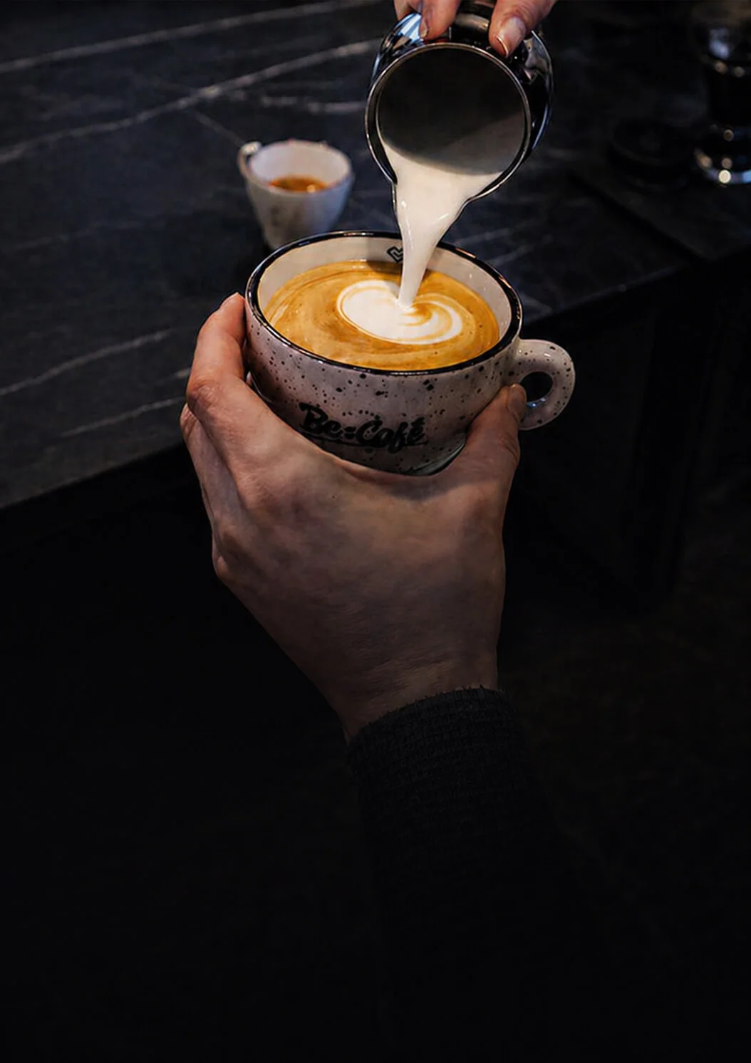 Barista nalieva napenené mlieko do kávy a vytvára latte art v keramickej šálke Be:Café